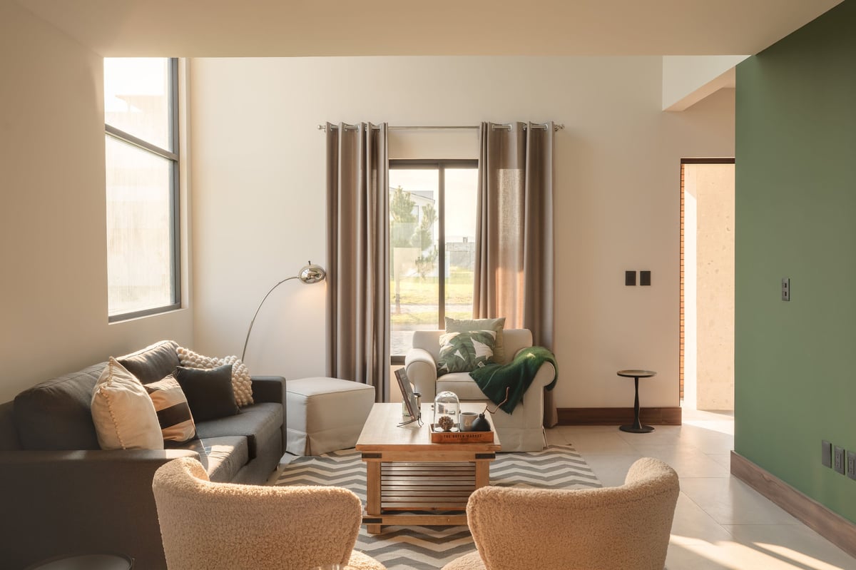 Modern living room with beige and green walls, gray sofa, two tan armchairs, wooden coffee table, and large windows with brown curtains